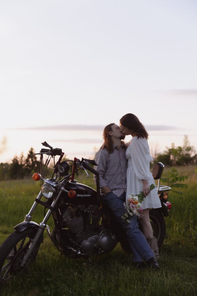 A couple leaning against a motorcycle in a field at sunset.